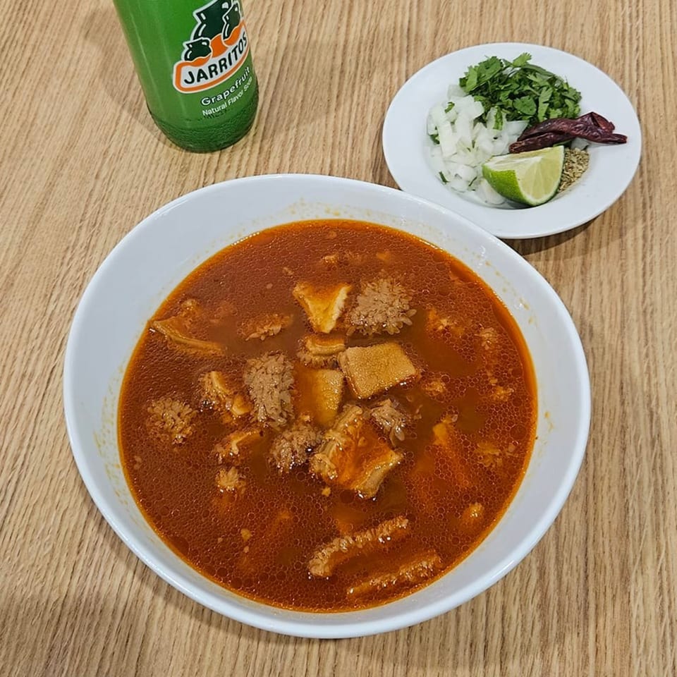 Menudo soup with a side of chopped onions, cilantro, lime, and dried chiles, with a Jarritos soda.