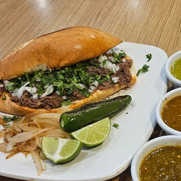 A Mexican torta with shredded meat, cilantro, and onions, served with grilled jalapeño, limes, and salsas.